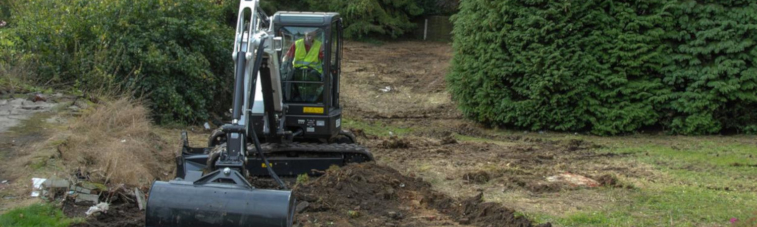 Person operating a white Bobcat® excavator while scooping up dirt with bushes and trees in the …